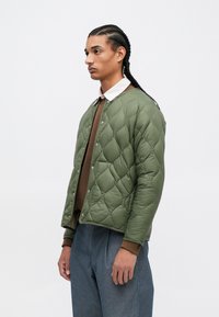 Jachetă verde matlasată cu capse, suprafață texturată și manșete ribbate, purtată peste o îmbrăcăminte maro, expusă pe un fundal simplu.