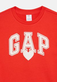 Κόκκινη βαμβακερή μπλούζα με κοντά μανίκια και λευκό λογότυπο "GAP" και σχέδιο καρδιάς στο μπροστινό μέρος. Στρογγυλή λαιμόκοψη, κοντά μανίκια και μαλακή υφή.