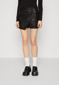 Mini-jupe en cuir noir avec une coupe asymétrique, ornée de deux accents en boucle en argent à la taille. Portée avec des chaussettes blanches et des chaussures noires.