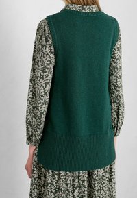 Grün gestrickte Weste mit geripptem Saum, getragen über einem langärmeligen Kleid mit floralem Muster in Creme- und Grüntönen.