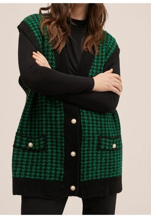 Vrouw draagt een groene en zwarte vest met pied-de-poule patroon en parelknopen over een zwart top met lange mouwen, armen gekruist.