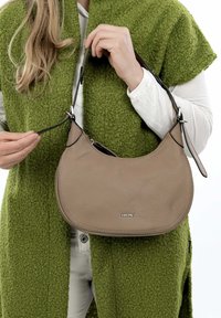 Borsa a tracolla in pelle marrone con forma arrotondata e superficie testurizzata. Presenta dettagli neri e una maniglia corta. Mantenuta elegante da un logo in argento.