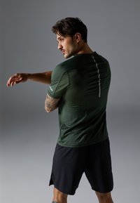 Green athletic shirt with short sleeves, featuring a slim fit and reflective accents. Paired with black shorts, showing a textured fabric.