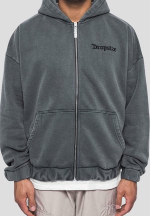 Homme portant un sweat à capuche gris zippé avec le texte « Dropsize » sur la poitrine, poches avant, et un pantalon beige avec une broderie « Dropsize » assortie.