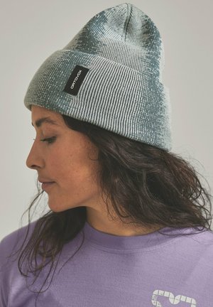 Woman with long dark hair wearing a gray knitted beanie labeled "ORTOVOX" and a light purple shirt, looking downward to the left.