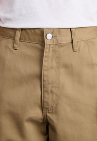 Tan cotton pants featuring a button closure, straight cut, and subtle texture, with visible side pockets and seam detailing.