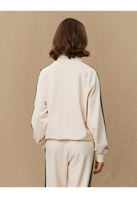 Cream-colored tracksuit featuring a plain design with green side stripes, made of soft fabric, presented from the back.