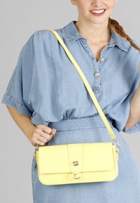 Borsa a tracolla gialla in pelle con finitura testurizzata, forma rettangolare, chiusura magnetica e tracolla regolabile, tenuta da una persona in un outfit di denim.