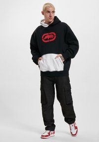 Zwarte hoodie met rode logo, witte kangoeroe zak en witte gevoerde kap, gecombineerd met zwarte cargo broek en rode sneakers.