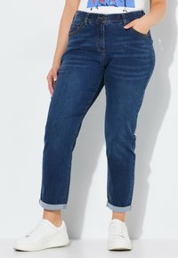 Jeans w ciemnym denimu o luźnym kroju, z podwiniętymi nogawkami, kieszeniami z przodu i subtelnymi detalami przeszyć. W zestawieniu z białymi trampkami.