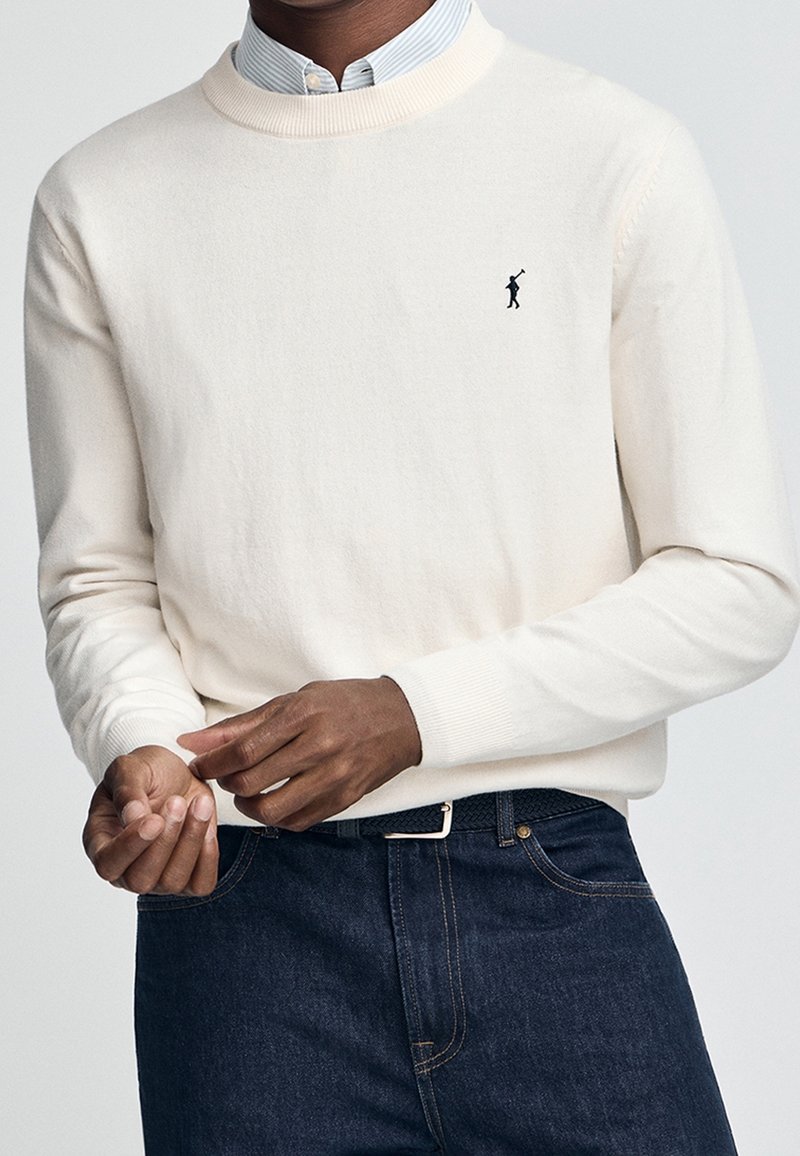 Homme portant un pull blanc à col rond avec un petit logo noir de joueur de polo, un jean bleu foncé et une chemise bleu clair à col en dessous.