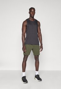 Débardeur noir sans manches associé à un short vert. Le mannequin porte des chaussures de sport noires et des chaussettes blanches avec un logo. Design simple et fonctionnel.