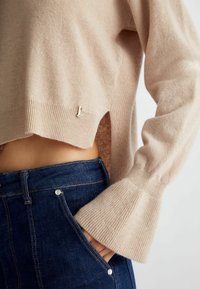 Maglione beige lavorato a maglia con polsini a coste e spacco laterale, abbinato a jeans in denim blu scuro con hardware argentato e vita aderente.