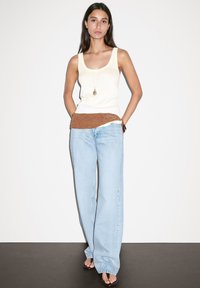 Top a fascia crema con accentuazione in crochet marrone, abbinato a jeans blu chiaro a gamba larga. Il modello sta in piedi con le mani sui fianchi contro uno sfondo neutro.