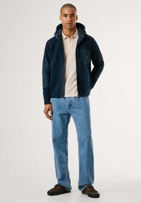 Hombre de pie con una chaqueta con capucha marina sobre una camiseta polo beige, jeans de color azul claro y zapatillas oscuras marrones, con un fondo sencillo.