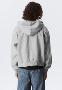 Personne aux cheveux bouclés portant un sweat à capuche gris et un jean bleu, debout de dos devant un fond uni.