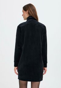 Zwarte corduroy jurk met een hoge kraag en lange mouwen, voorzien van verticale ribstructuur en een aansluitende silhoeët.