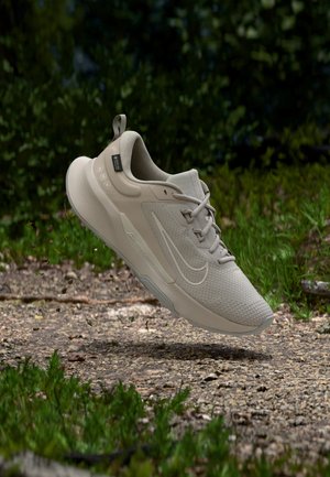 Chaussure de sport de course beige avec lacets et logo, en l'air au-dessus d'un chemin de gravier avec de la verdure en arrière-plan.