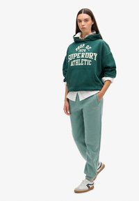 Sudadera verde corta con texto blanco, combinada con pantalones de chándal de color verde azulado claro. La modelo lleva zapatillas blancas con detalles en negro y marrón.