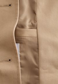 Close-up of beige fabric with stitched buttonholes and a partially visible white label inside a garment seam.