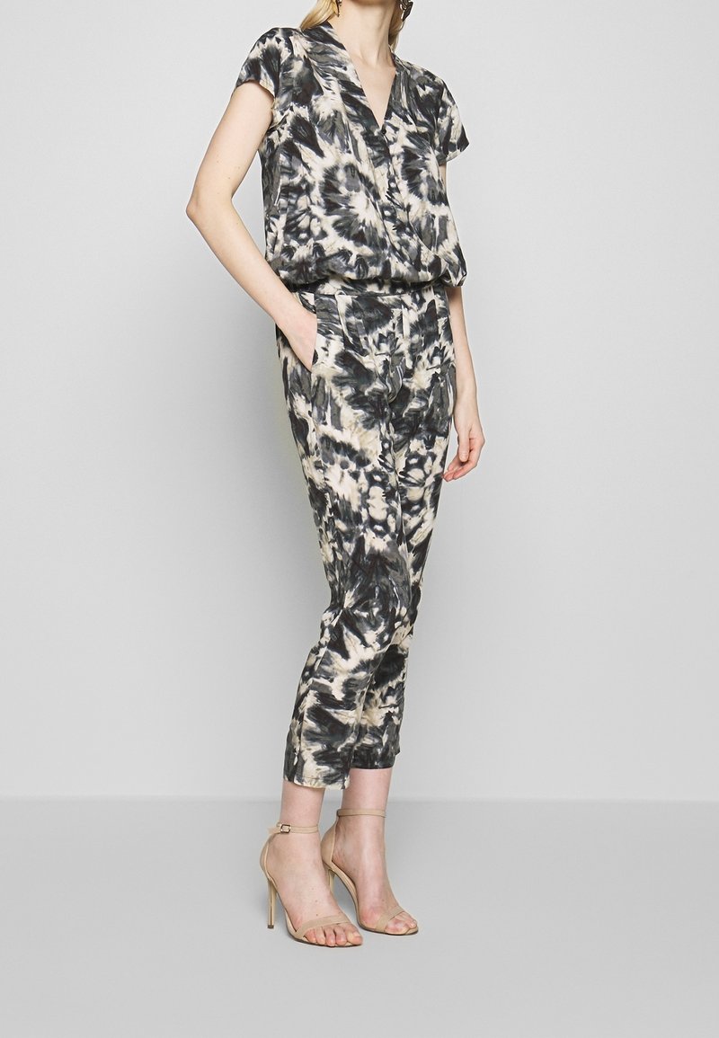 Femme portant un chemisier à manches courtes tie-dye noir et beige et un pantalon court assorti, avec des sandales à talons hauts beige, debout.