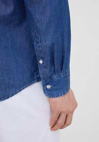 Camicia di denim con una texture morbida; presenta un colore blu scuro, maniche lunghe e polsini con bottoni con un leggero dettaglio di arricciatura.