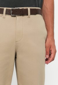 Beige cotton trousers with a smooth texture, featuring a brown leather belt with a silver metal buckle, and slanted side pockets.