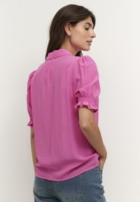 Pink blouse with puffed short sleeves, pleated collar, and soft fabric. Worn with jeans, showing a relaxed fit and smooth texture.