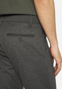 Pantalon gris en tissu texturé, doté d'une poche arrière avec fermeture à bouton et d'une petite étiquette de marque attachée sur le côté.