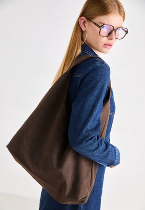 Jeune femme portant de grandes lunettes écaille et des boucles d’oreilles créoles porte un grand sac marron sur une veste en jean bleue.