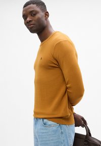 Musztardowy sweter z długim rękawem o okrągłym dekolcie, ze ściągaczami przy rękawach i na dole; noszony z jasnoniebieskimi dżinsami i brązową torebką.