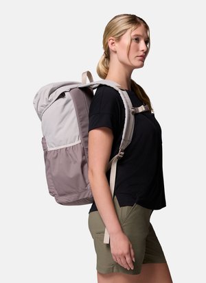 Ung kvinde i sort skjorte og grønne shorts iført en stor tofarvet grå vandrerygsæk, stående sidelæns på hvid baggrund.