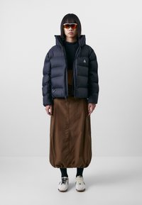 Black puffer jacket with a hood, ribbed black sweater, and brown skirt. White athletic shoes and sunglasses with red-tinted lenses.