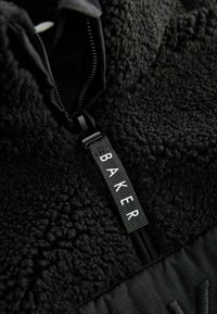 Zwarte fleece jas met een ritskraag. Kenmerkt zich door textiel met structuur, een soepele rits en een label met de tekst "BAKER." Contrasterende panelen aan de onderkant.
