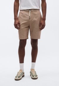 Μπεζ λινά σορτς με ελαστική μέση και κορδόνι. Συνδυασμένα με πράσινα και μπεζ αθλητικά παπούτσια, λευκές κάλτσες και ένα απλό λευκό T-shirt.