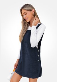 Vestido de tirantes de pana de color marino con tirantes ajustables, botones laterales y un corte holgado, usado sobre una blusa ajustada de manga larga blanca.