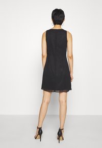 Robe noire sans manches, longueur genou, tissu lisse avec une légère brillance, fermeture éclair au dos, accentuée par un ourlet transparent, associée à des chaussures à talons hauts.