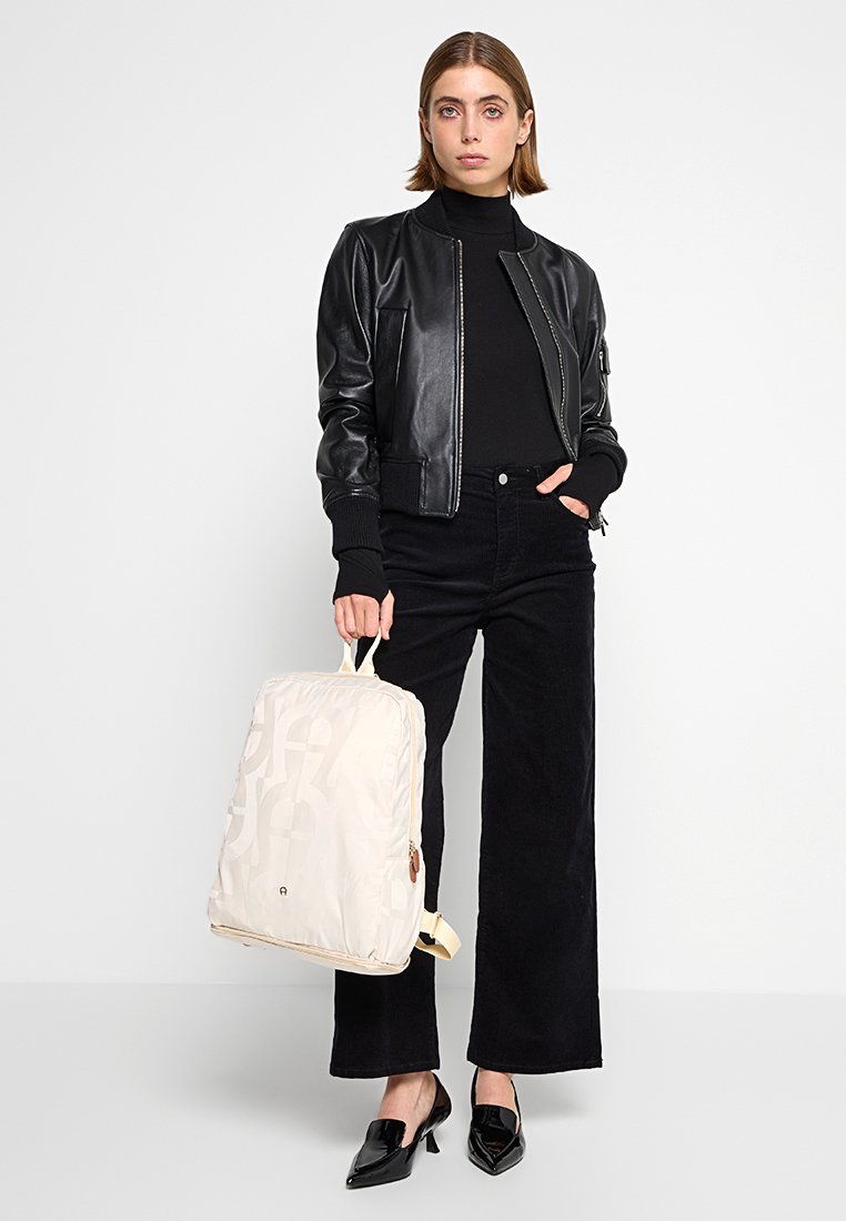 Schwarze Lederbomberjacke, schwarzes Rollkragenshirt und weite schwarze Jeans. Hellbeige Rucksack mit subtilen Mustern und Reißverschlussdetails.