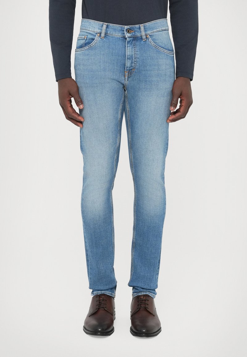 Jeans de ganga azul claro com corte justo, com cinco bolsos, passadores de cinto e detalhes de costura visíveis. Usados com sapatos castanho escuro.