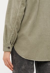 Chemise en velours côtelé vert olive avec un ourlet arrondi, des poignets boutonnés et une texture côtelée, associée à un pantalon texturé sombre.