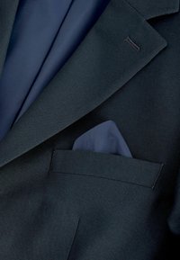 Veste de costume bleu foncé avec un mouchoir de poche bleu assorti plié dans la poche poitrine sur un fond sombre.