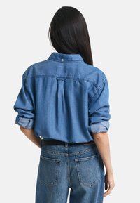 Denim shirt in light blue with a button-down collar, rolled sleeves, and a back pleat. Paired with medium-wash straight-leg jeans.