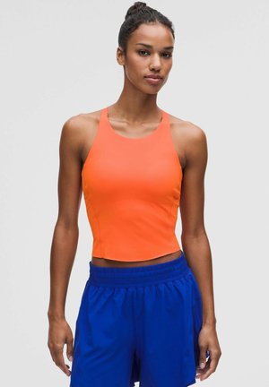 Kvinde iført en orange ærmeløs cropped top og kongeblå shorts med elastik i taljen står mod en ensartet lys baggrund.