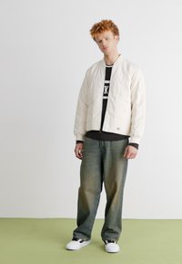 Cremefarvet bomberjakke med kviltet tekstur, parret med en mørk langærmet skjorte og løstsiddende, slidte jeans. Hvide sneakers fuldender looket.