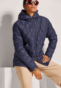 Marineblauer, gesteppter Pufferjacke mit Kapuze, Reißverschluss und strukturiertem Stoff. Modell in beigen Hosen und übergroßen Sonnenbrillen.