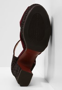 Chaussure à talon bloc à bride arrière avec semelle noire embossée et empeigne foncée à motif, reposant sur un coin de sol et mur blancs.