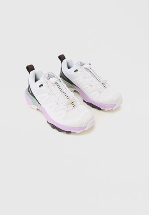 Een paar witte sportschoenen met roze tussenzolen, zwarte en groene accenten, en elastische veters op een lichte, effen achtergrond.