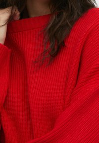 Personne aux longs cheveux foncés portant un pull rouge texturé, tenant le col du pull près de son visage avec une main.