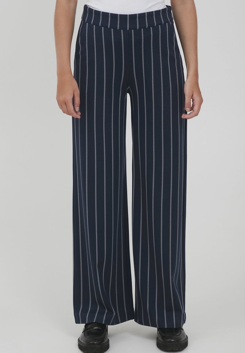 Personne portant un pantalon large marine à rayures verticales blanches et des chaussures noires, debout devant un fond gris uni.