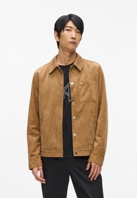 Suede rjava jakna s ovratnikom, gumbi na pritisni in žepom na prsih. Podedovana z črno srajco z grafičnim potiskom.